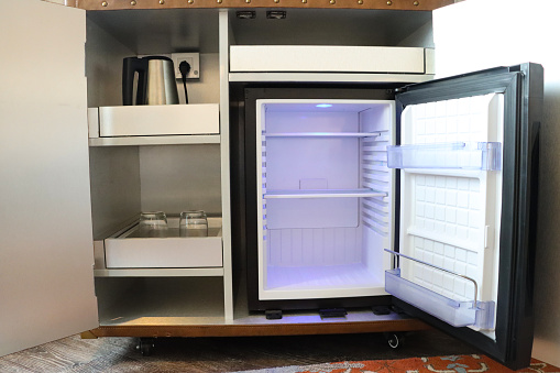 Stock photo showing close-up view of hotel cabinet with integrated fridge with door opened to display shelves and interior light and shelves with electric kettle and drinking glasses.