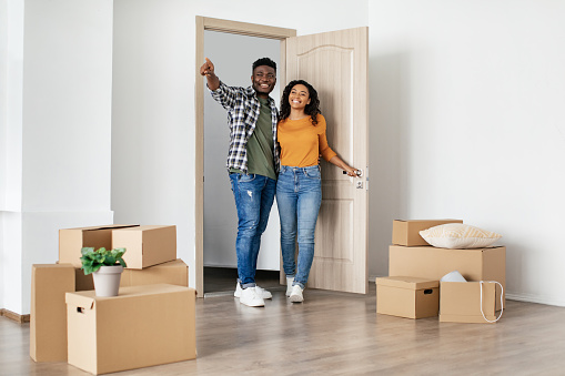 Real Estate Ownership. African American Husband And Wife Entering Their New House Standing In Living Room Among Moving Boxes At Home. Apartment Rent And Purchase. Full Length Shot