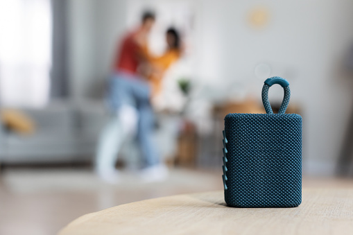 Selective focus on brand new gadget portable speaker over dancing romantic lovers, blurred background, copy space, home interior. Modern technologies and domestic entertainment concept