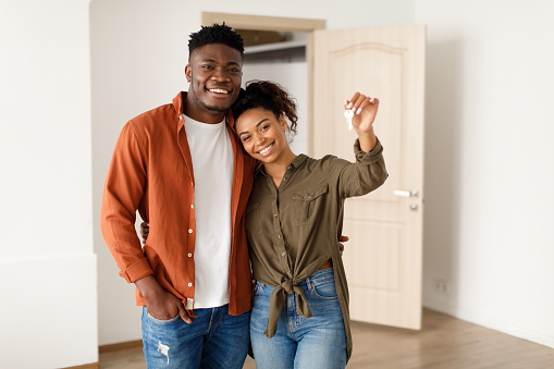 Real Estate. Cheerful African American Spouses Showing New House Key Smiling To Camera, Embracing Standing Among Moving Cardboard Boxes At Home. Relocation, Apartment Ownership And Family Housing