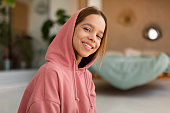 Portrait of cheerful caucasian teen girl wearing hood and smiling at camera, posing sitting in bedroom interior at home