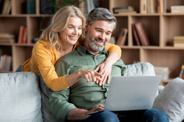 Happy Middle Aged Couple With Laptop Ordering Things From Internet Together Happy Middle Aged Couple With Laptop Ordering Things From Internet Together While Relaxing On Couch In Living Room, Smiling Mature Spouses Making Virtual Shopping Or Booking Hotel Online, Closeup mature couple stock pictures, royalty-free photos & images