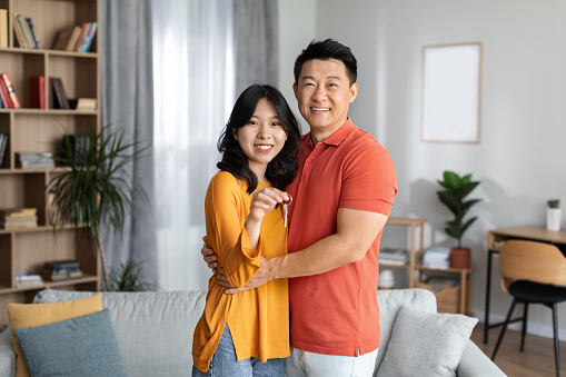 Happy asian couple in casual outfits hugging in the living room, showing key from their new apartment and smiling at camera, enjoying new house, copy space. Mortgage, house loan concept