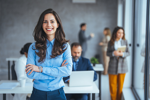 Successful businesswoman standing in creative office and looking at camera. Young woman entrepreneur in a coworking space smiling. Portrait of beautiful business woman standing in front of business team at modern agency with copy space.