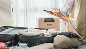 Closeup of young Asian muslim woman preparing suitcase use smartphone checking list accessories in bedroom at home. Travel vacation.