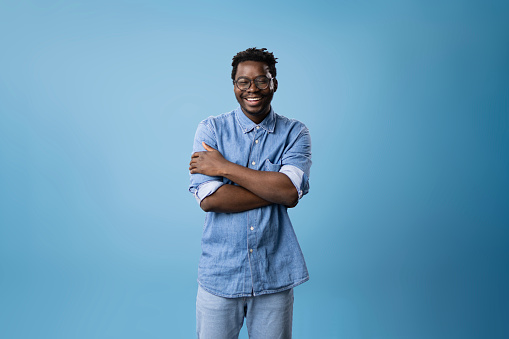 Studio shot of happy African American man in jeans standing against blue background with his arms crossed and looking at camera. Copy space.