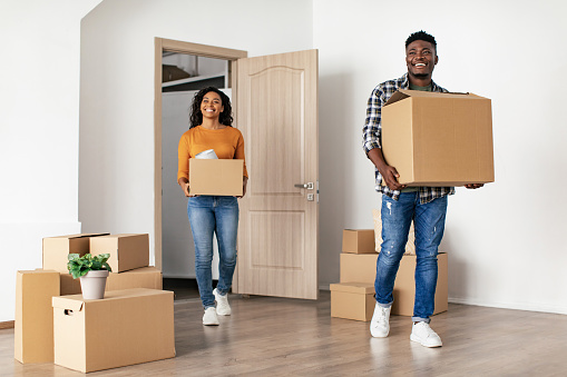 New Home. Black Couple Entering New Apartment Carrying Cardboard Boxes Moving House Posing Indoor. Family Looking At Empty Living Room. Relocation, Real Estate Offer Concept