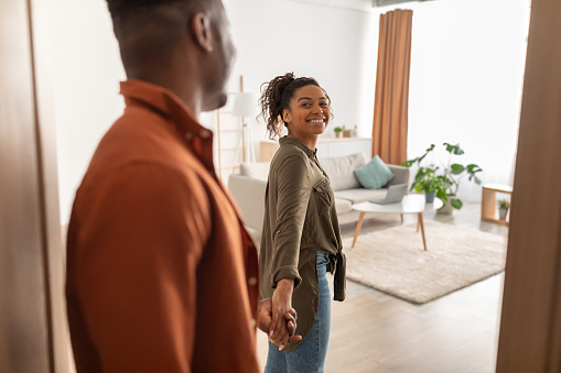 Welcome To New Home. Happy African American Couple Holding Hands Standing In Opened Doors Entering Their Apartment Indoors. Real Estate Ownership. Selective Focus On Female