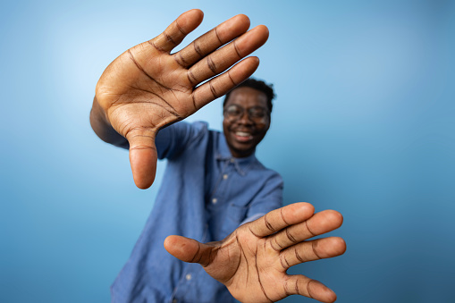 Close up of African American man making a photo frame with his hands against blue background. Copy space.