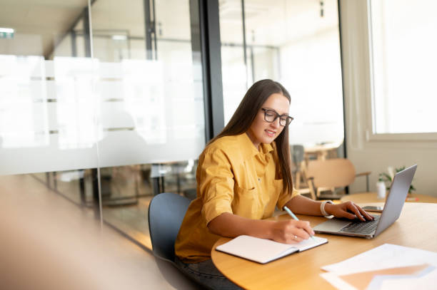 istock Smiling female student using laptop for watching webinars, lectures, sits at desk in office and taking notes, woman taking a part in online educational course 1469156263