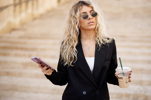 Young businesswoman in black suit walking down the stairs, drinking take away coffee and texting on smart phone outdoors