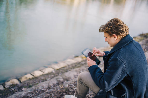 istock Front view of young man using digital tablet outside in the park next to river. Copy space. stock photo 1469157121
