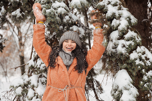Attractive young woman in a warm pink coat having fun, smiling and trotting a snow-covered branch and snow is falling on it. Winter entertainment