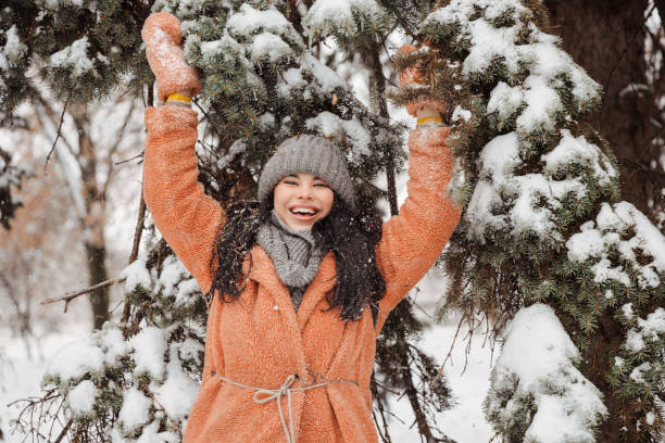 Attractive young woman in a warm pink coat having fun, smiling and trotting a snow-covered branch and snow is falling on it. Winter entertainment stock photo