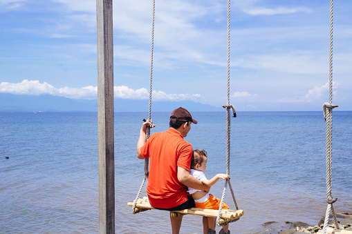 father and baby boy swinging on a big beautiful wooden swing on the beach -