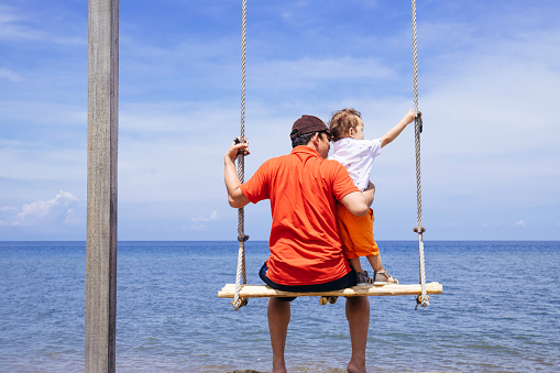 father and baby boy swinging on a big beautiful wooden swing on the beach -