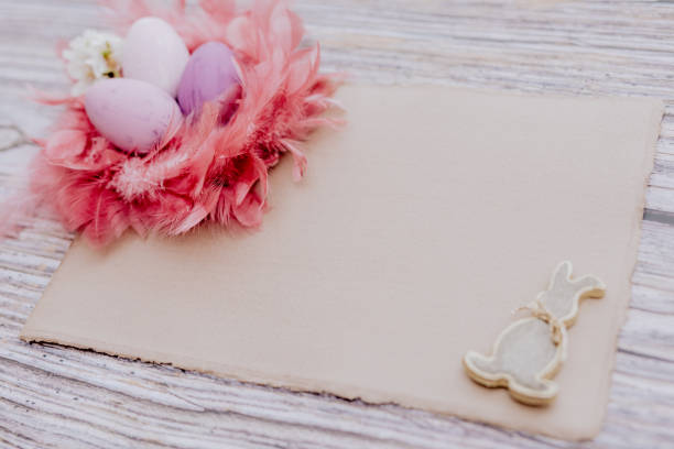 Close up of a mockup consisting of an empty beige handcraft DNA4 paper, different purple Easter eggs in a pink feather nest and an Easter bunny shape on a rustic wooden background with copy space. Color editing with added grain. Very selective and soft focus. Part of a series.