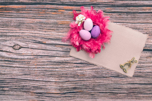 Mockup flat lay consisting of an empty beige handcraft DNA4 paper, different purple Easter eggs in a pink feather nest and a metallic painted Easter bunny on a rustic wooden background with copy space. Color editing with added grain. Very selective and soft focus. Part of a series.