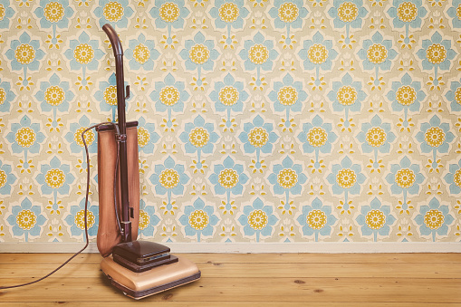 Retro styled image of a vintage brown vacuum cleaner in a room with floral wallpaper and wooden floor