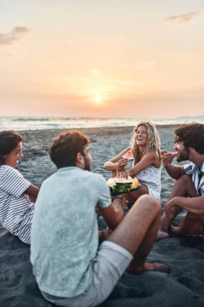 istock Cheerful couples talking while eating fruit on the beach at sunset. 1469167429