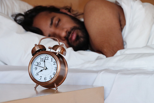 Stock photo showing close-up view of a bronze metal case retro alarm clock with double bell on night stand. An Indian man can be seen lying in the double bed, next to the beside table, sleeping.