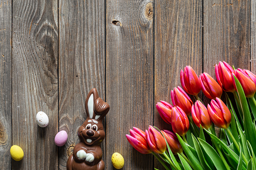 Easter background with a bouquet of pink tulips, a chocolate bunny and Easter eggs on a wooden background, top view, copy space.