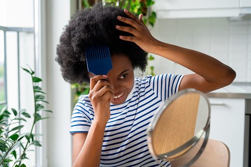Cool African woman with comb sticking out of curly hair smiling looks in mirror and touches wig with hands. Positive black teen girl with wavy hairstyle grooms herself sits at table in home