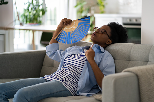Tired exhausted African American woman sits on sofa at home using paper fans and closing eyes suffering from stuffiness. Tormented black girl housewife is resting after hot walk in summer weather
