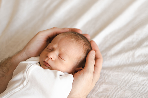 Father,young man holding caucasian hairy brunet cute newborn baby sleeping.One or two week child on bed in male hands indoors.Care, love, happiness concept. Copy space for text.