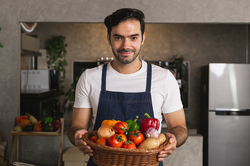 Close up Caucasian man wearing apron hand holding fresh organic vegetable tomato bell pepper potato garlic onion basket on wooden table in home kitchen. Young man chef select good quality ingredient.