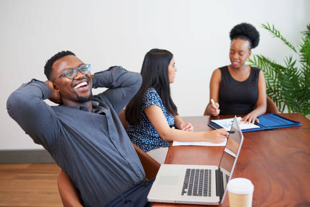 Black businessman laughs and relaxes at boardroom table arms behind head
