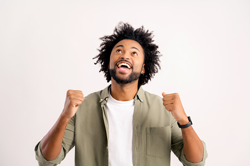 Excited African-American curly guy raising hands in ecstatic standing isolated on white, lucky man won in lottery, celebrating success, looking up and screaming yes, triumph and victory concept