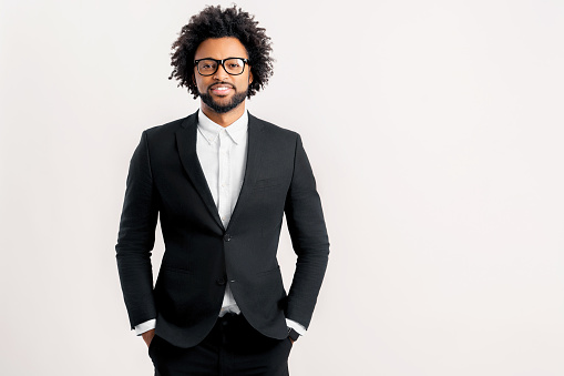 Smiling curly businessman standing with arms in pockets, looking at camera isolated on white. Young positive male entrepreneur in black formal suit. Proud successful multiracial small business owner