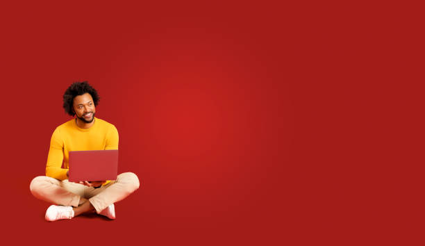 istock Confident handsome african american curly man sitting on the floor in lotus pose and using laptop isolated on red 1469199781