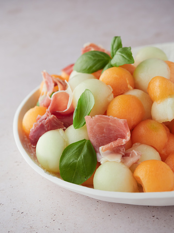 fruit salad with two varieties and colors of melon balls, serrano ham and fresh basil leaves. close up view half of a white plate.