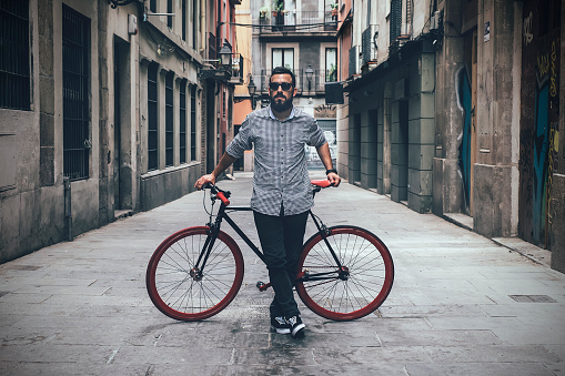 Full length of modern male in casual outfit and sunglasses leaning on bicycle while standing on empty narrow town street and looking at camera