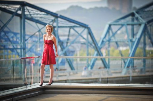 A girl in a red dress stands, holding onto the glass rail behind her and smiling, in front of the blue Walnut Street Bridge in Chattanooga TN.