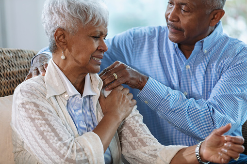 A senior African-American woman sitting at home on a couch, complaining about pain radiating from her shoulder down to her hand. Her husband tries to provide pain relief by massaging her shoulder.