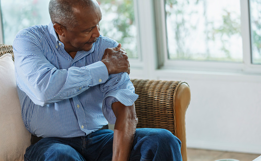 A senior African-American man sitting at home on a couch, rubbing his sore shoulder. It is painful from arthritis or an injury.