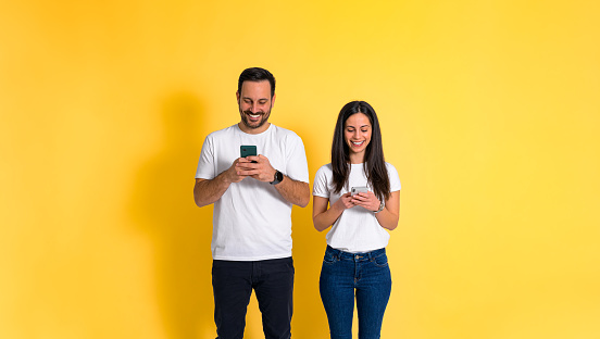 Happy young couple wearing white t-shirts chatting over smart phones while standing isolated against yellow background