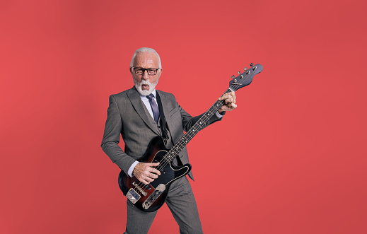 Portrait of excited senior bearded businessman singing cheerfully and playing electric guitar isolated over red background