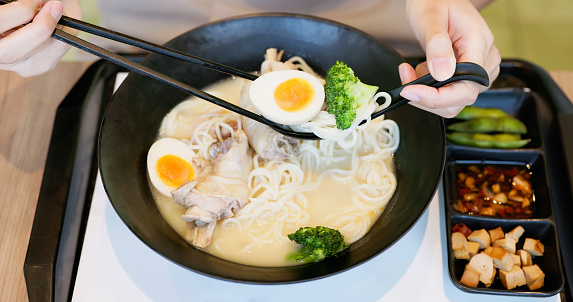 close up asian woman hand hold chopsticks and spoon eating tasty hot steaming japanese ramen soup noodle with meat and eggs on table in food court or restaurant