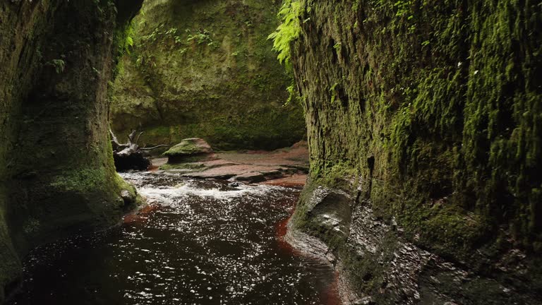 Drone shot creeping through the Devils Pulpit gorge in Scotland revealing mossy walls and a flowing red river.