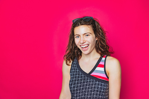 Portrait of beautiful young woman with sunglasses in brown wavy hair, wearing colorful shirt and smiling excitedly at camera, magenta background, studio shot