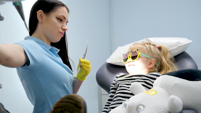 pediatric dentistry young beautiful doctor turns on light showing girl tools with which to treat teeth caries color directs to girl's face on sunglasses in hands a soft toy the first trip to dentist