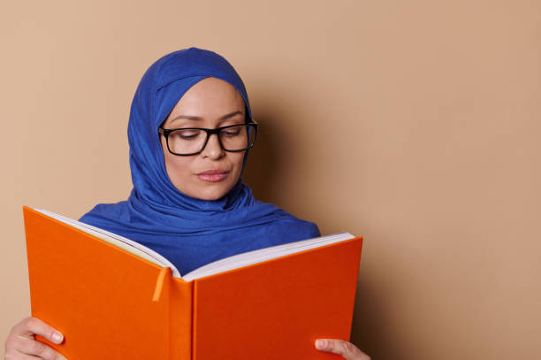 istock Educated sophisticated Muslim woman smiling at camera while reading book, standing isolated on a beige cream background 1469240305