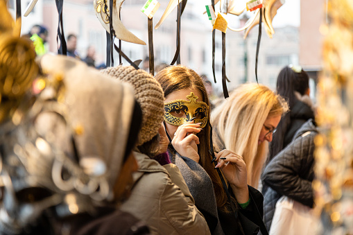 Venice, Veneto, Italy - Feb 19, 2023: Young woman tries a mask at a souvenir street store during 2023 Venice carnival celebrations