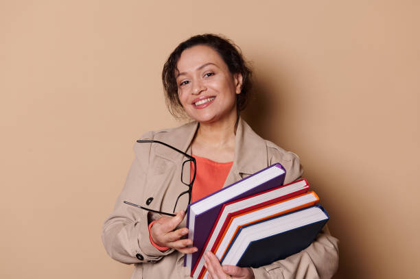 istock Middle-aged woman teacher at a high school institution, smiling at camera, carrying a stack of books on beige background 1469241776