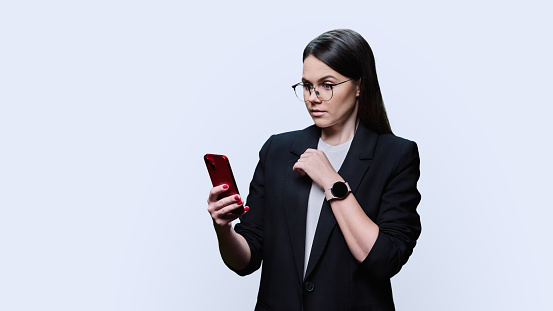 Young business woman using smartphone on white studio background. Positive smiling female in glasses jacket looking texting on phone. Mobile applications, digital technologies in business work leisure