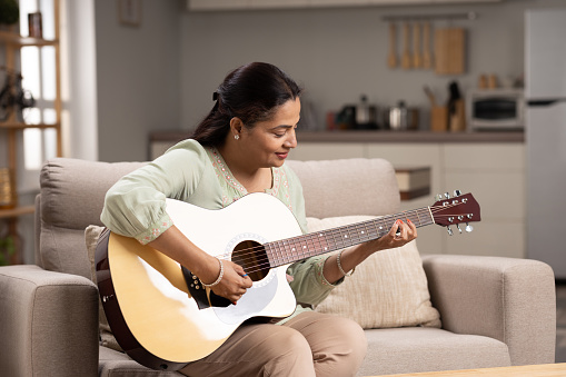 Mature woman in casual clothes is smiling while playing guitar at home